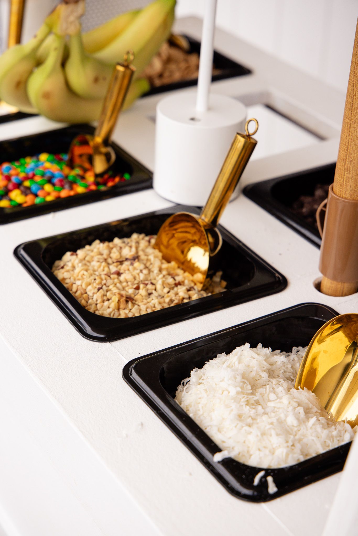 Various toppings and ingredients displayed in black serving trays with gold scoops for a dessert or snack bar setup.
