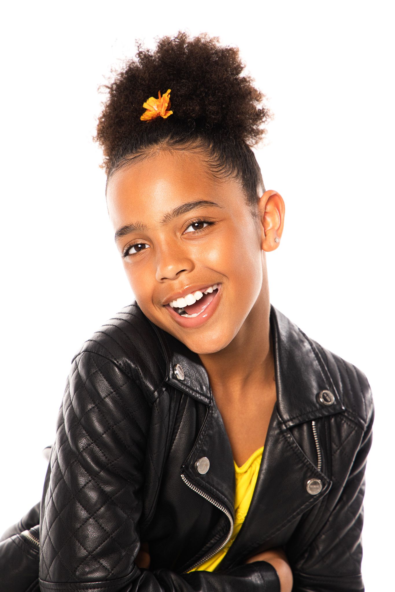 Smiling person wearing a black leather jacket and yellow top with curly hair in updo and orange flower accessory.
