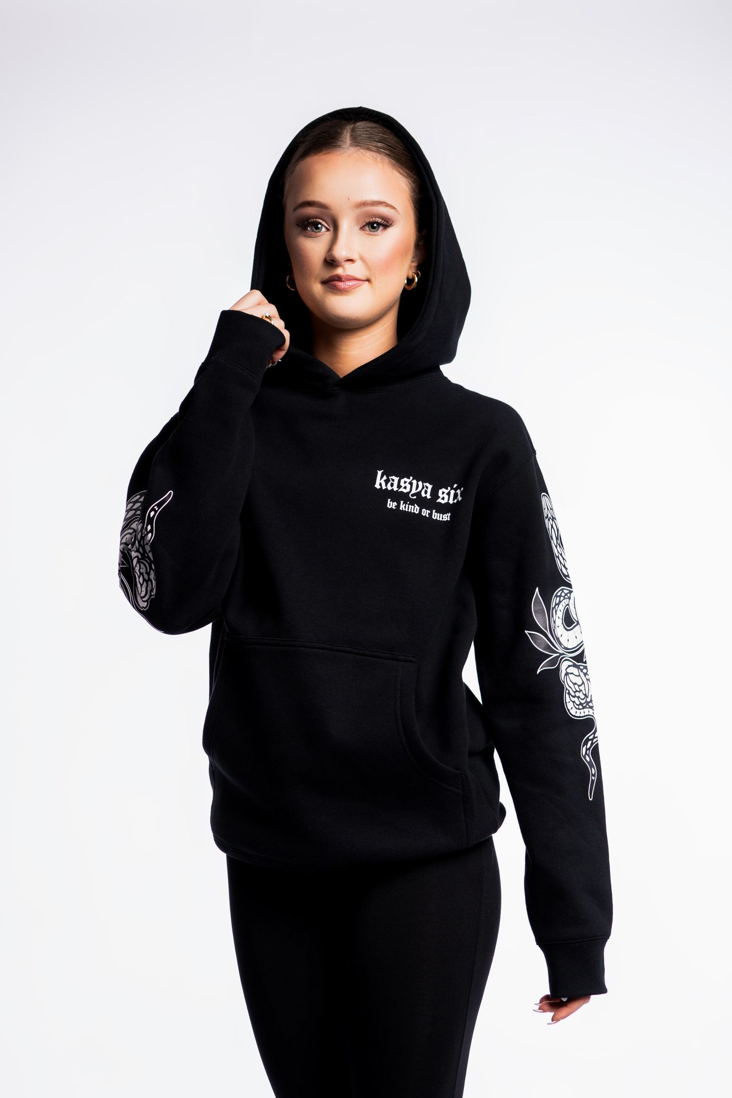 Woman wearing black hoodie with white graphic sleeve print and front logo, paired with black leggings.