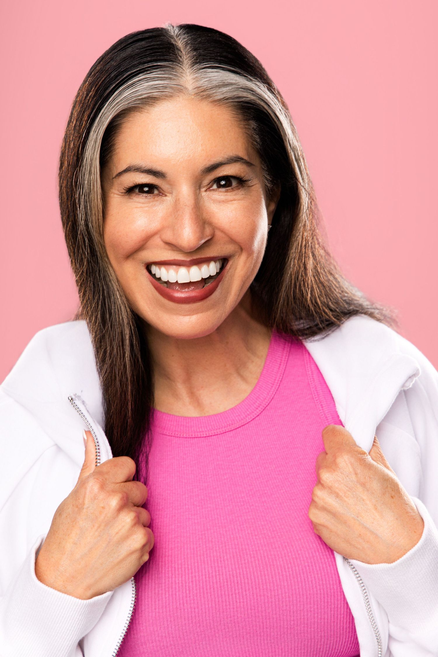 Smiling middle-aged woman with gray-streaked hair wearing a pink top and white zip-up jacket against a pink background.