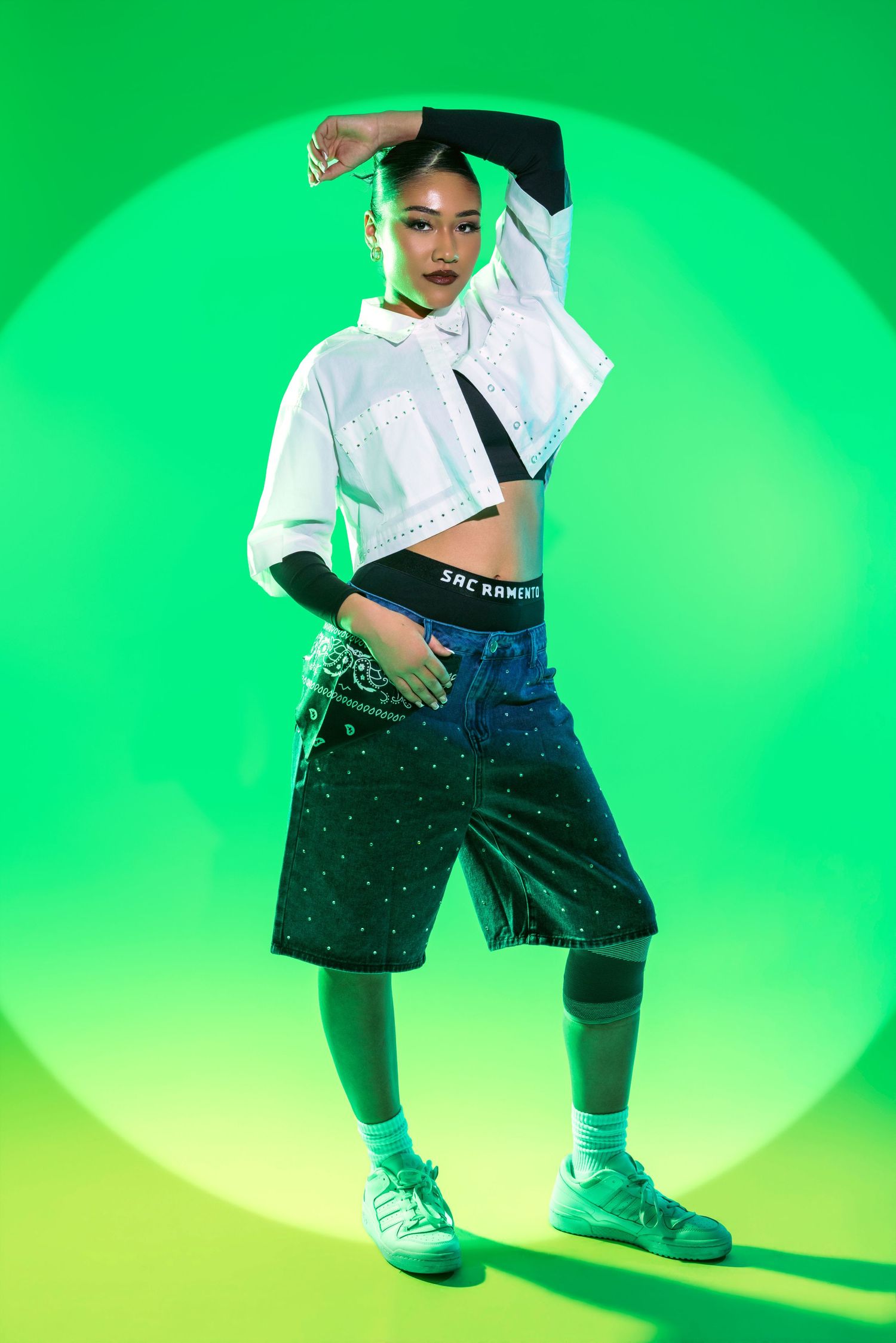Young woman in white crop top and dark shorts poses confidently against vibrant green background in streetwear fashion shoot.