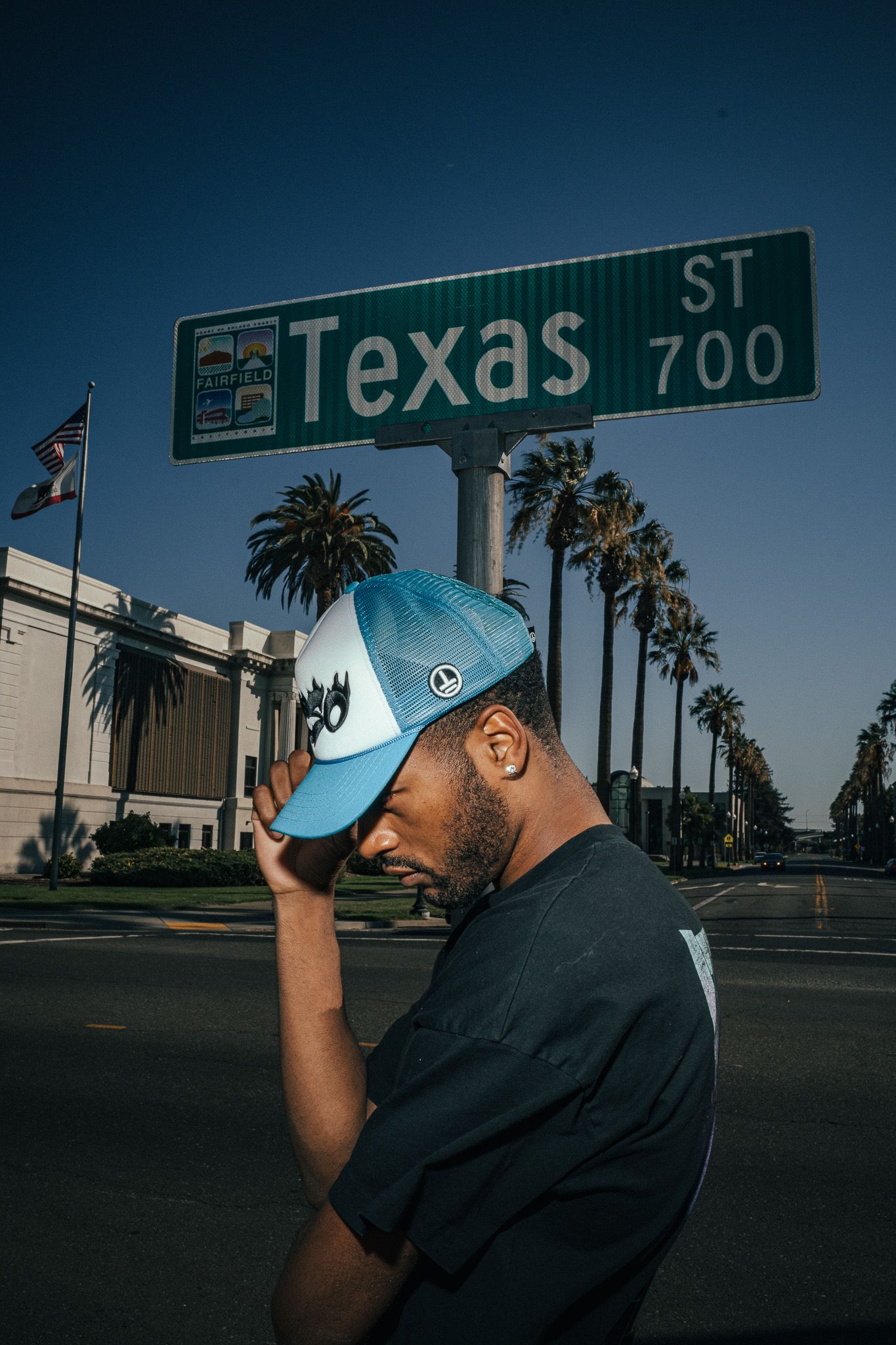 Man in blue cap stands under Texas St 700 sign with palm trees and blue sky in background.