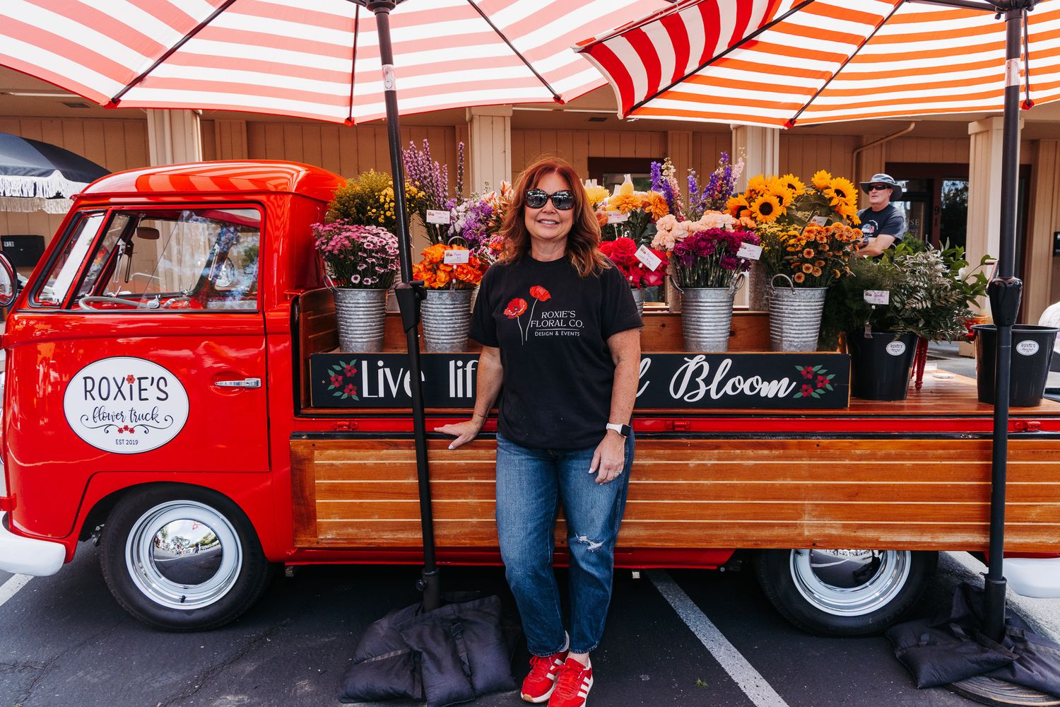 Woman poses beside red vintage VW flower truck with colorful blooms at outdoor market.