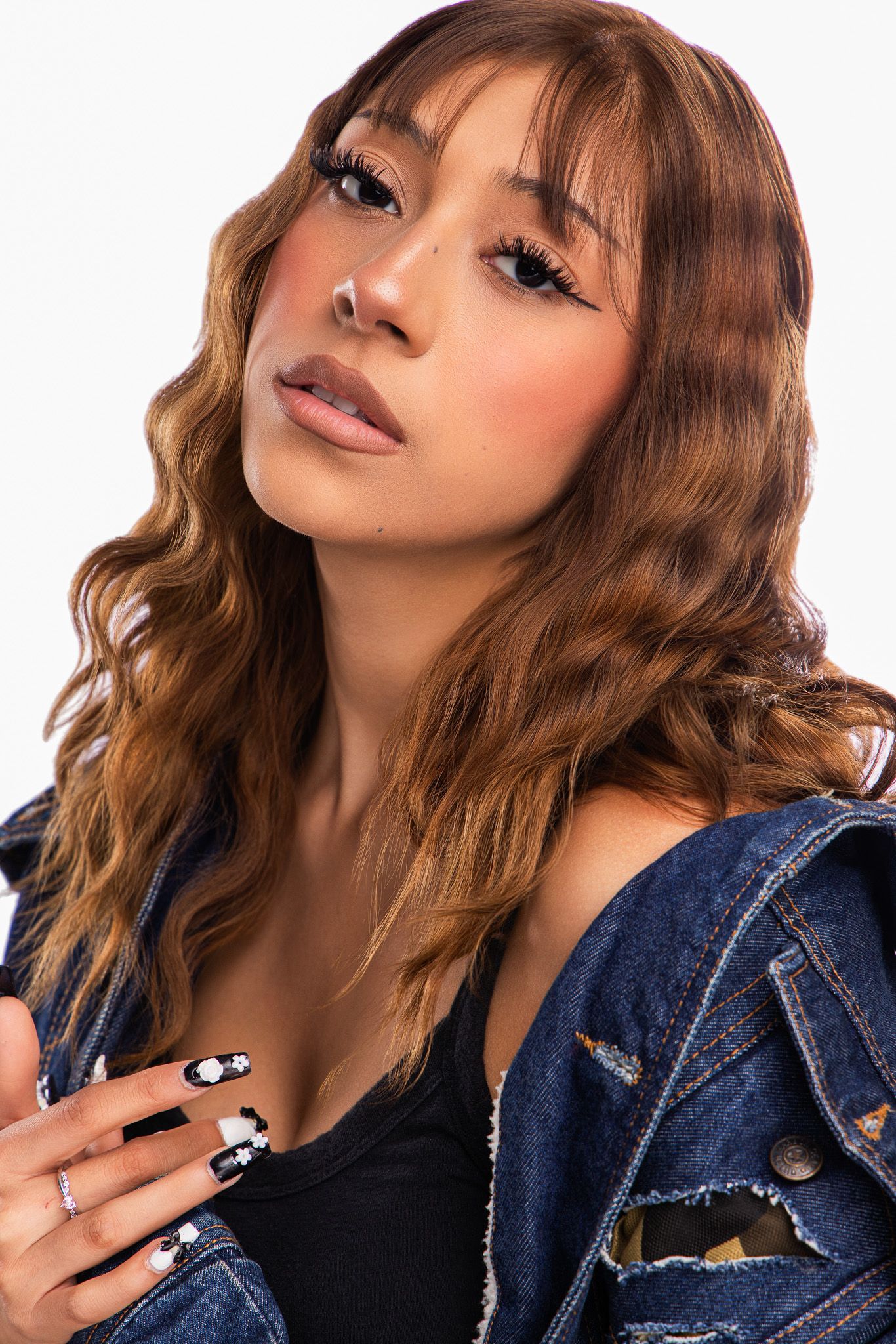 Young woman with auburn wavy hair, nose ring, black nail art, wearing denim jacket over black top, posing against white background.