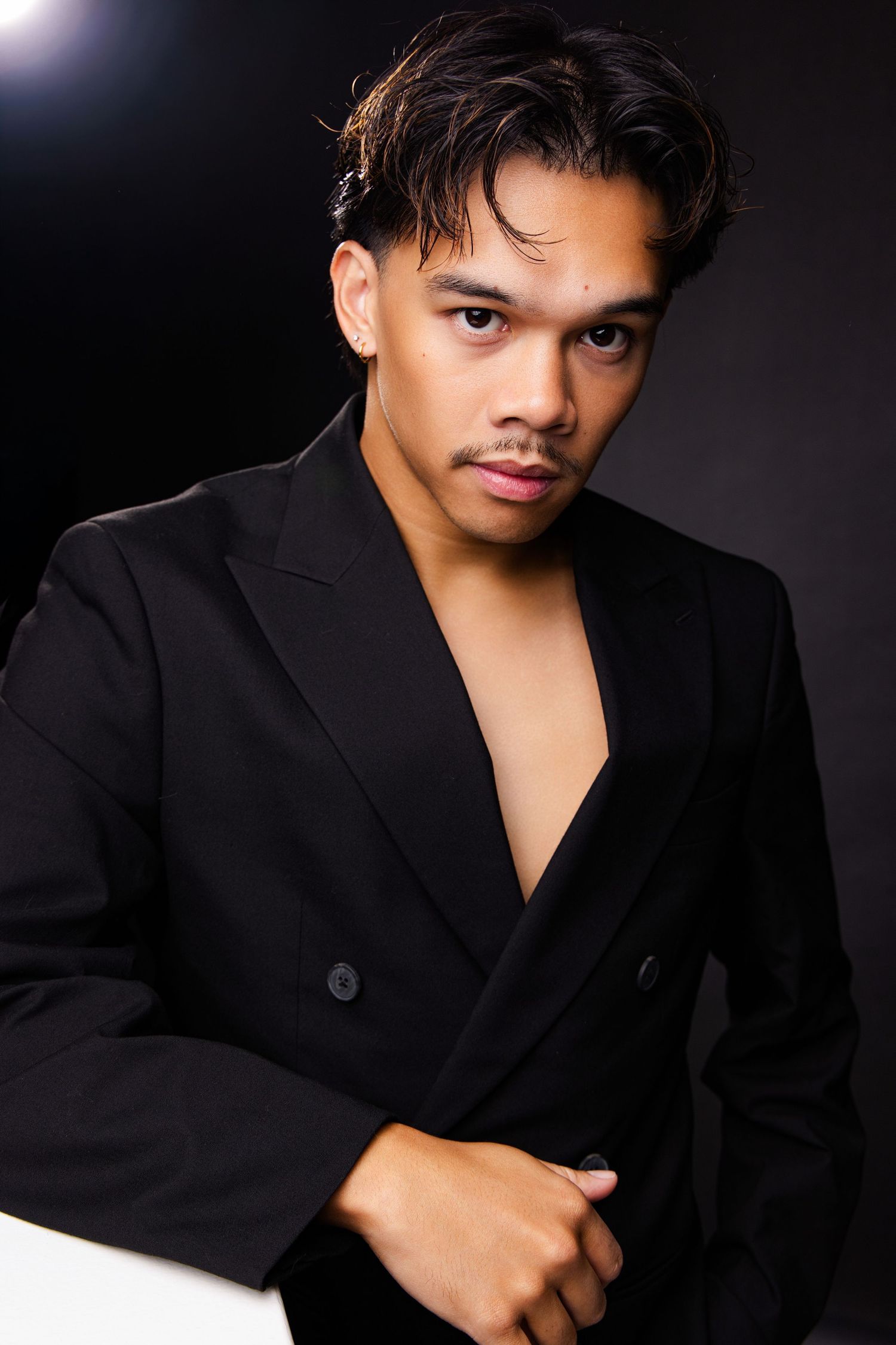 Young man in black blazer with open chest poses confidently against dark background in stylish fashion portrait.