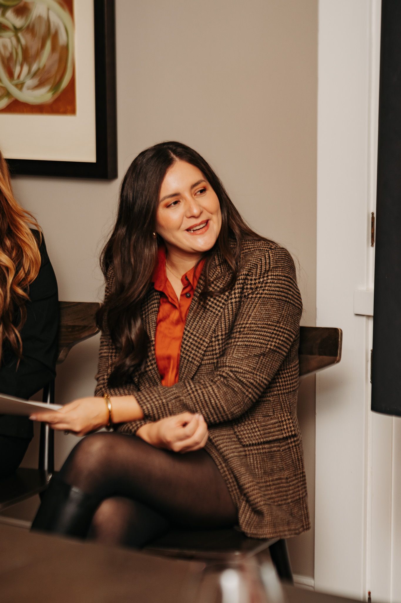 Person in tweed jacket and red top laughing while relaxing in an indoor setting.