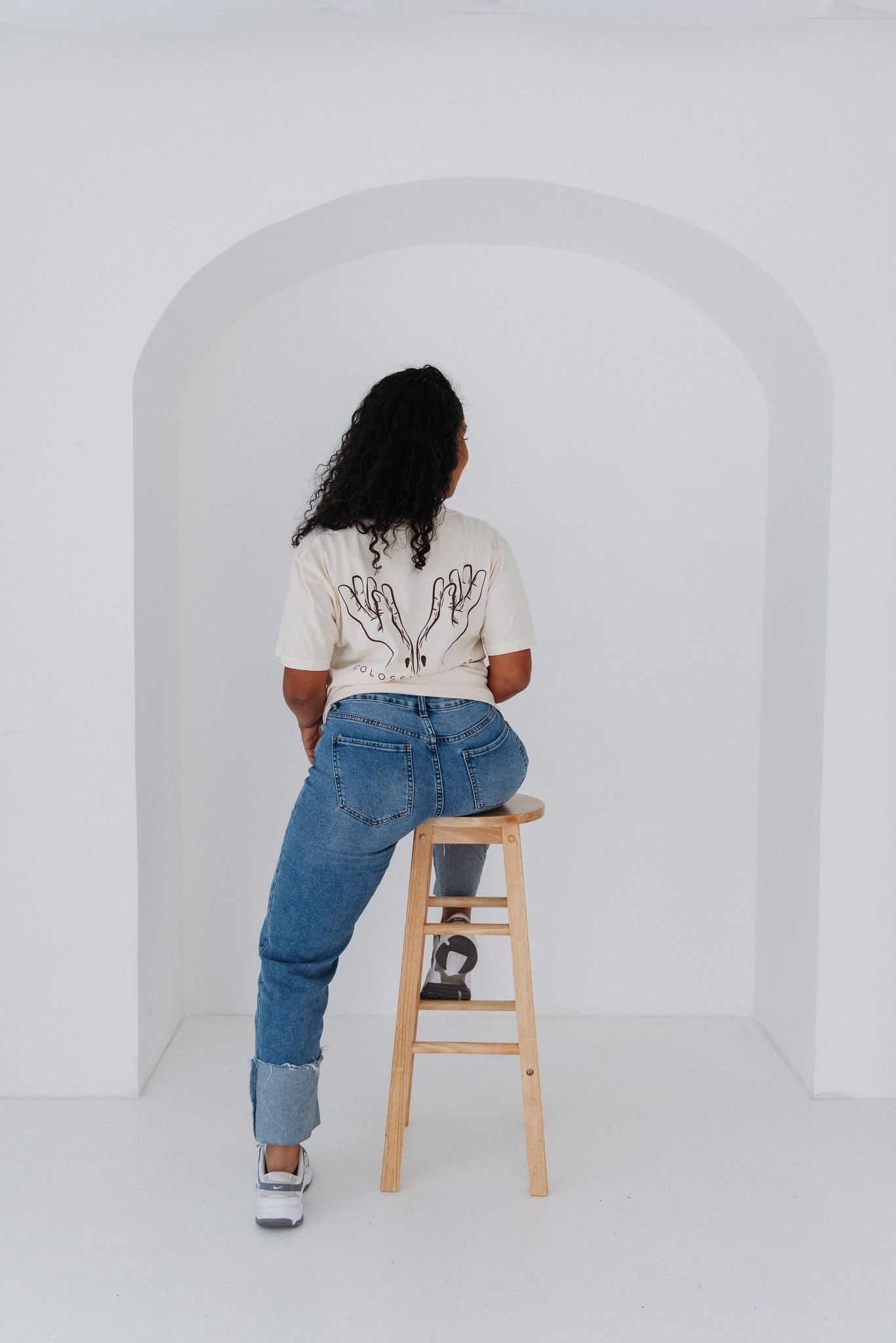 Person in white tee and rolled up jeans sits on wooden stool against minimalist white archway.
