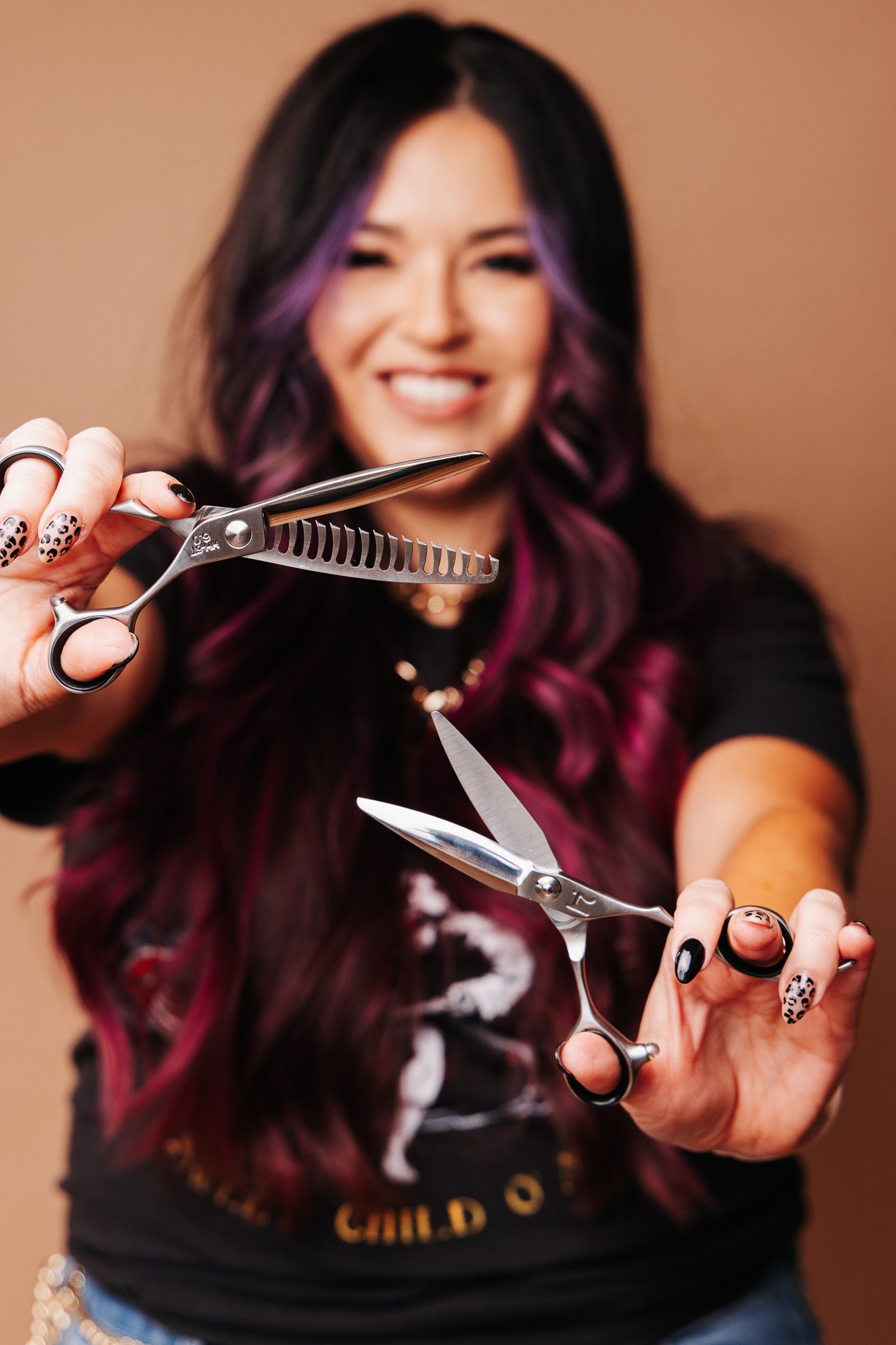 A person with purple-tinted hair displays hairstyling scissors against a brown studio background.