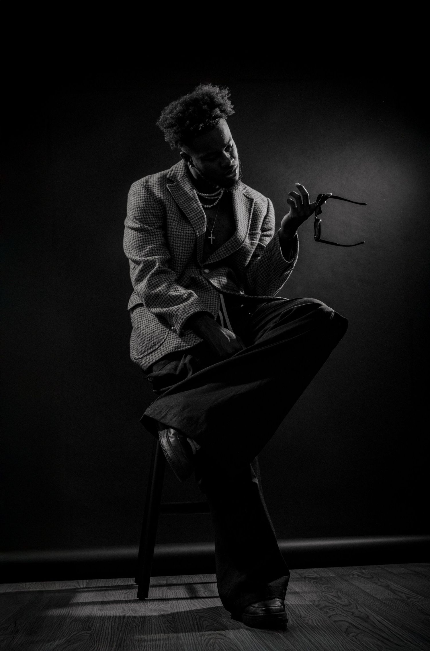 Dramatic black and white portrait of a performer holding a microphone while seated on a stool against dark background.