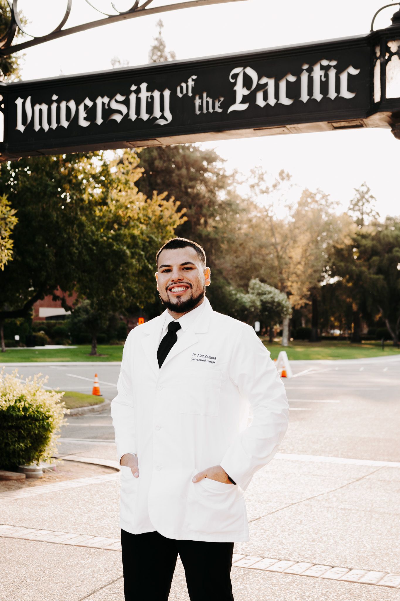 A person in a white coat stands smiling on a University of the Pacific campus pathway on a sunny day.