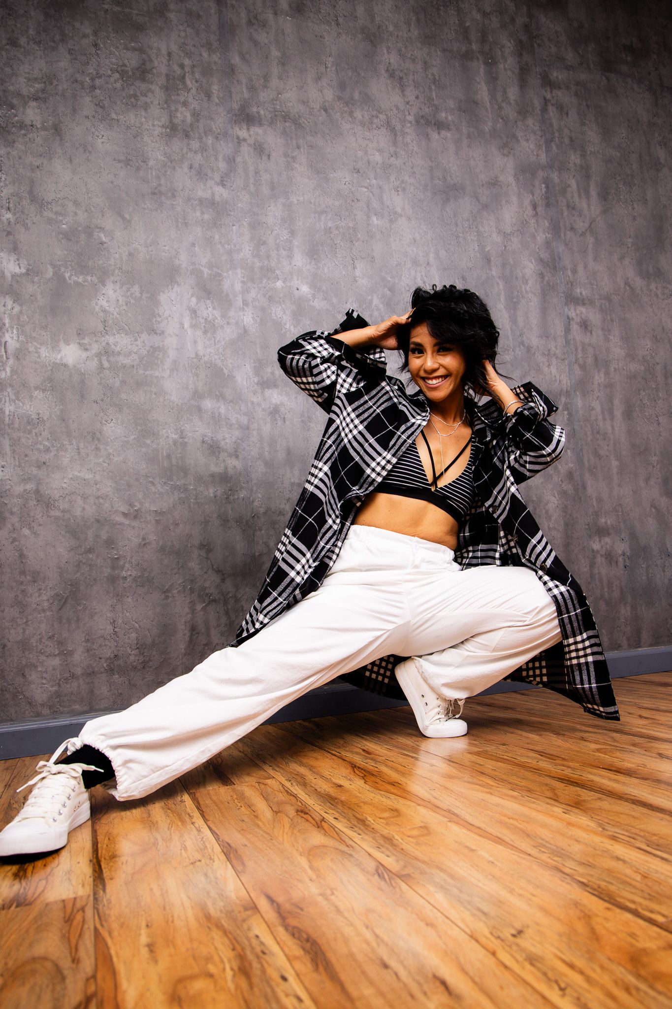 Urban dancer in plaid shirt and white pants performs dynamic floor moves against grey wall.