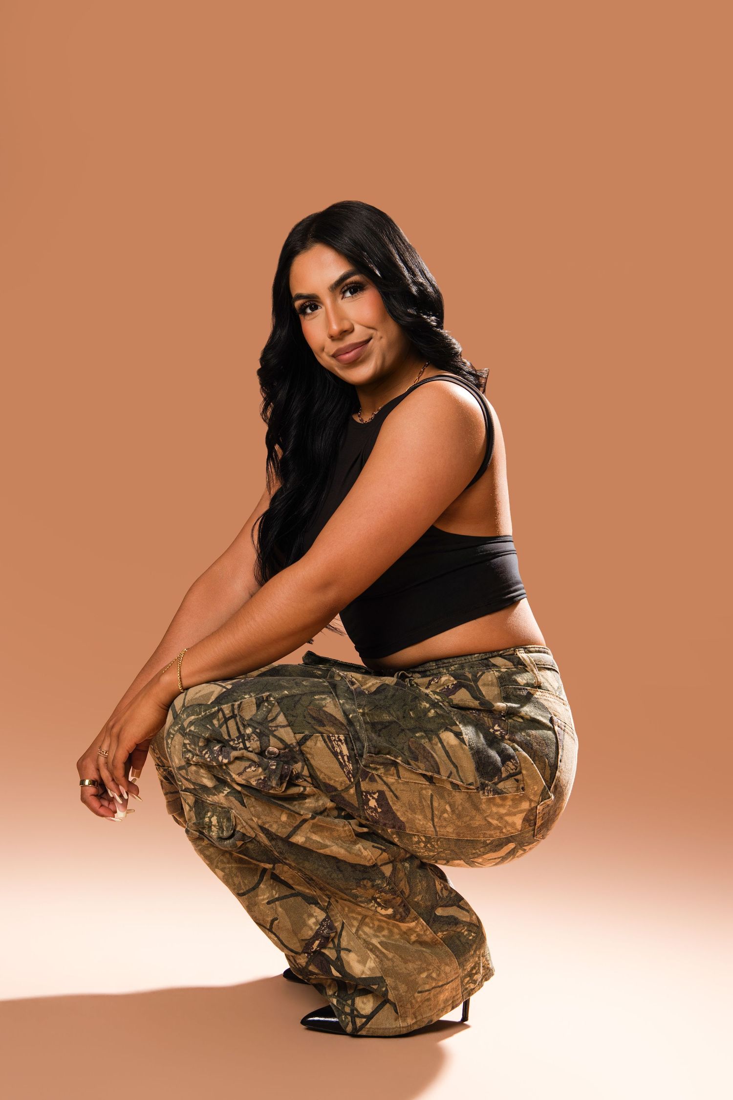 Woman in black crop top and camo cargo pants crouching, posing confidently against a warm brown background.