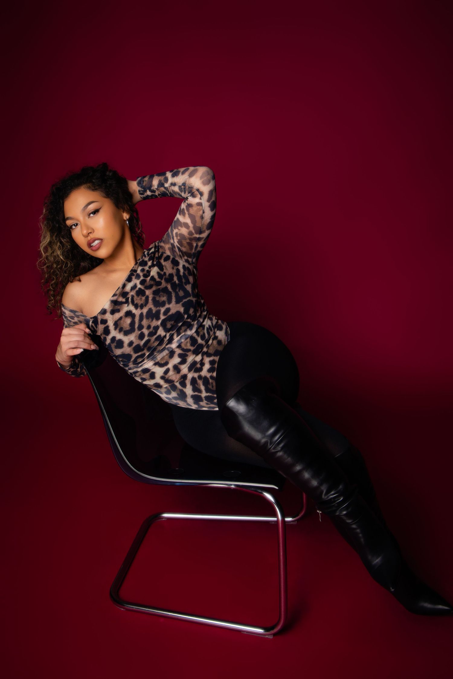 Model poses on modern chrome chair wearing leopard print top and black pants against dark burgundy backdrop.