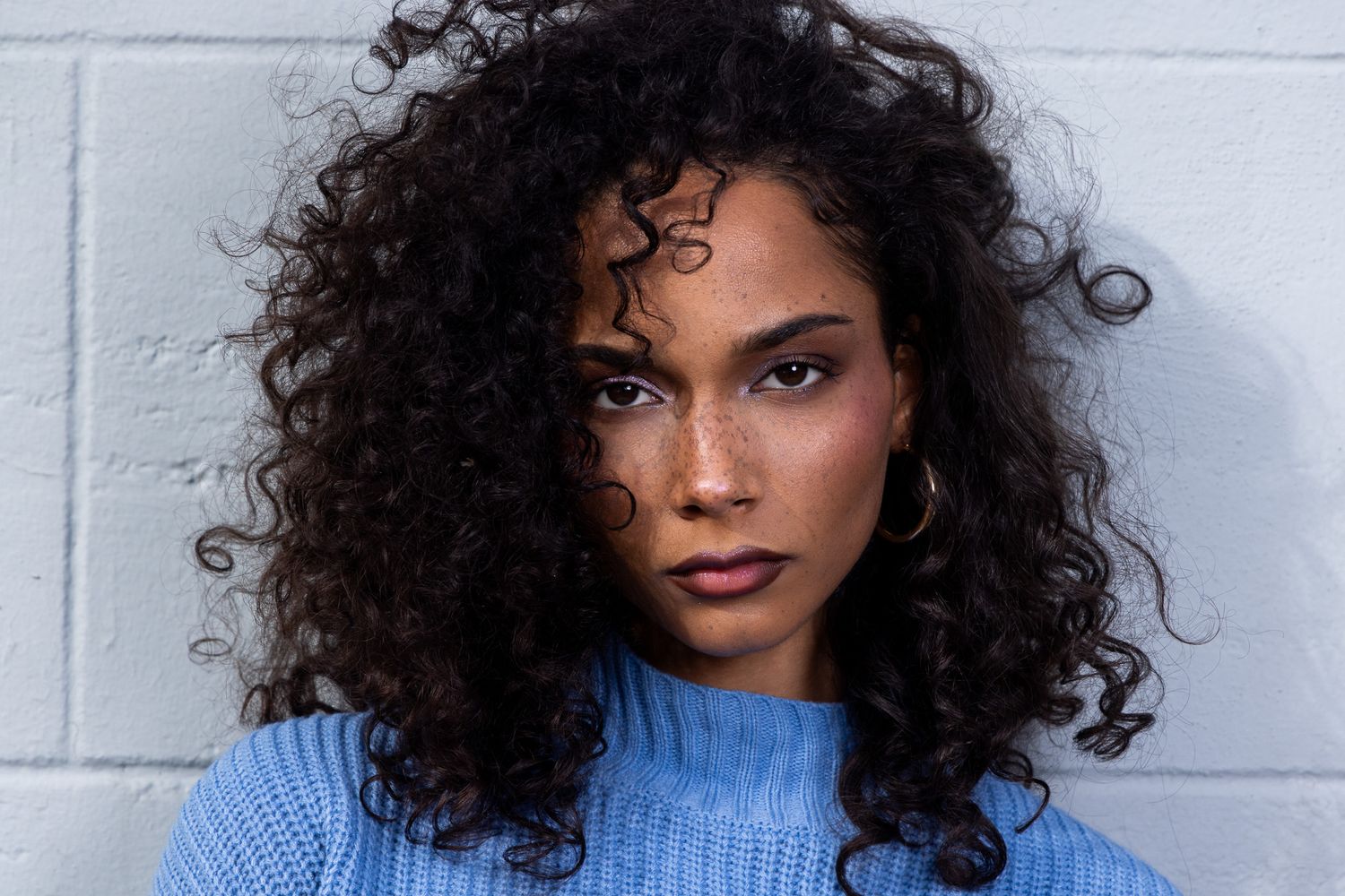 Side profile poses in light blue sweater against white brick wall.