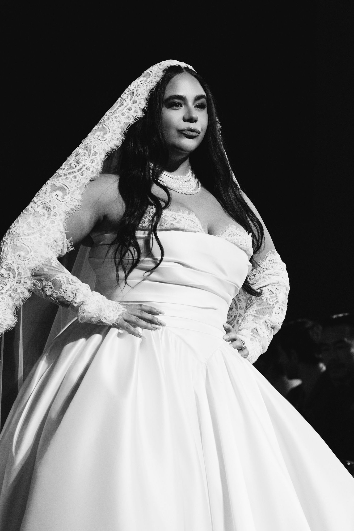 Black and white fashion photograph showing a dramatic white wedding gown with lace sleeves against dark background.