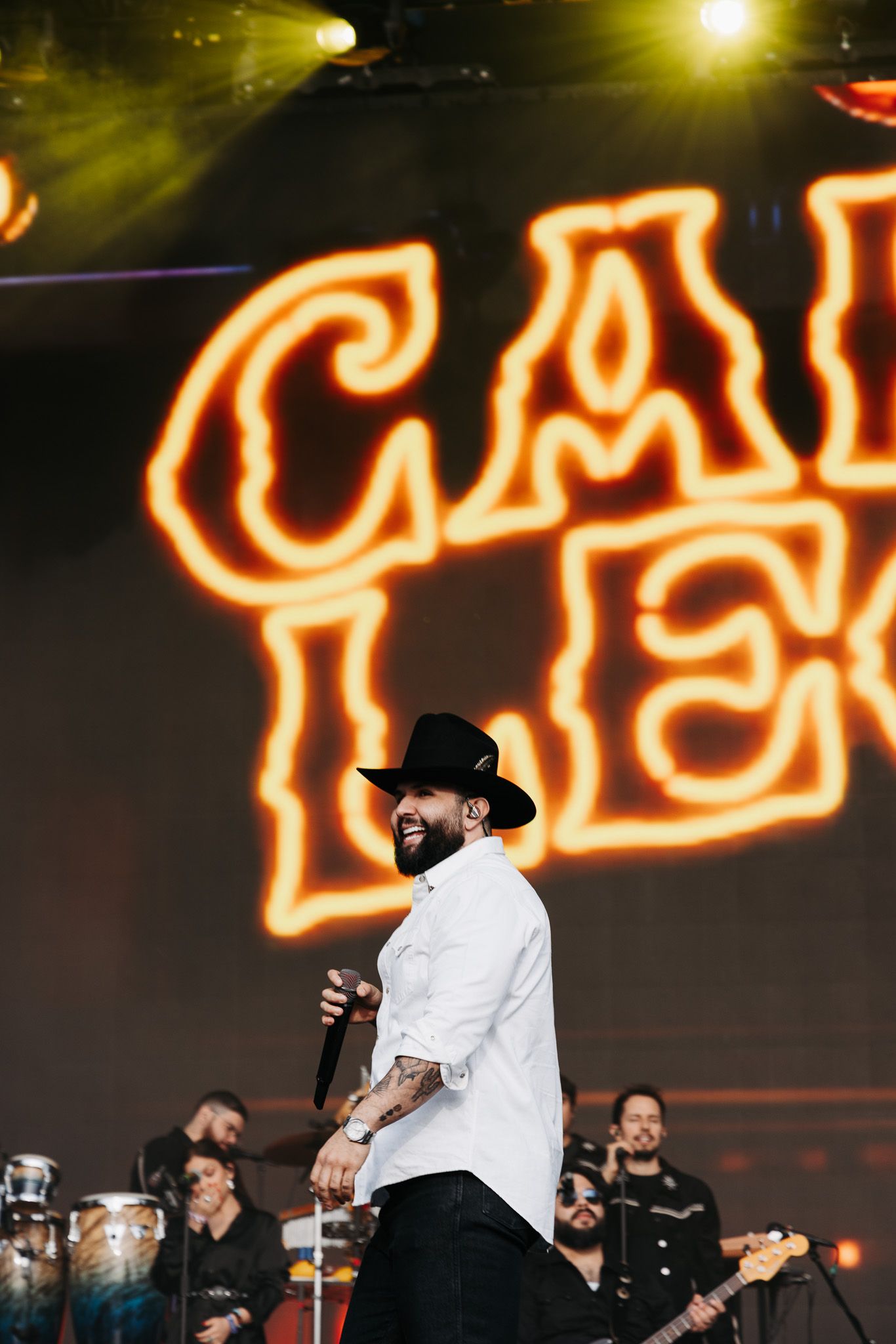 Concert performer in white shirt and black hat on stage with bright orange neon signage in background.