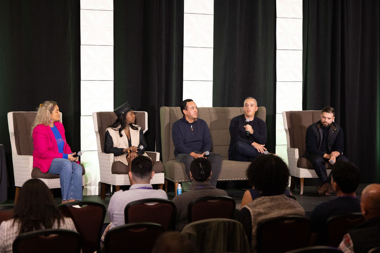 A panel of speakers sit in chairs on a stage during a professional conference event.