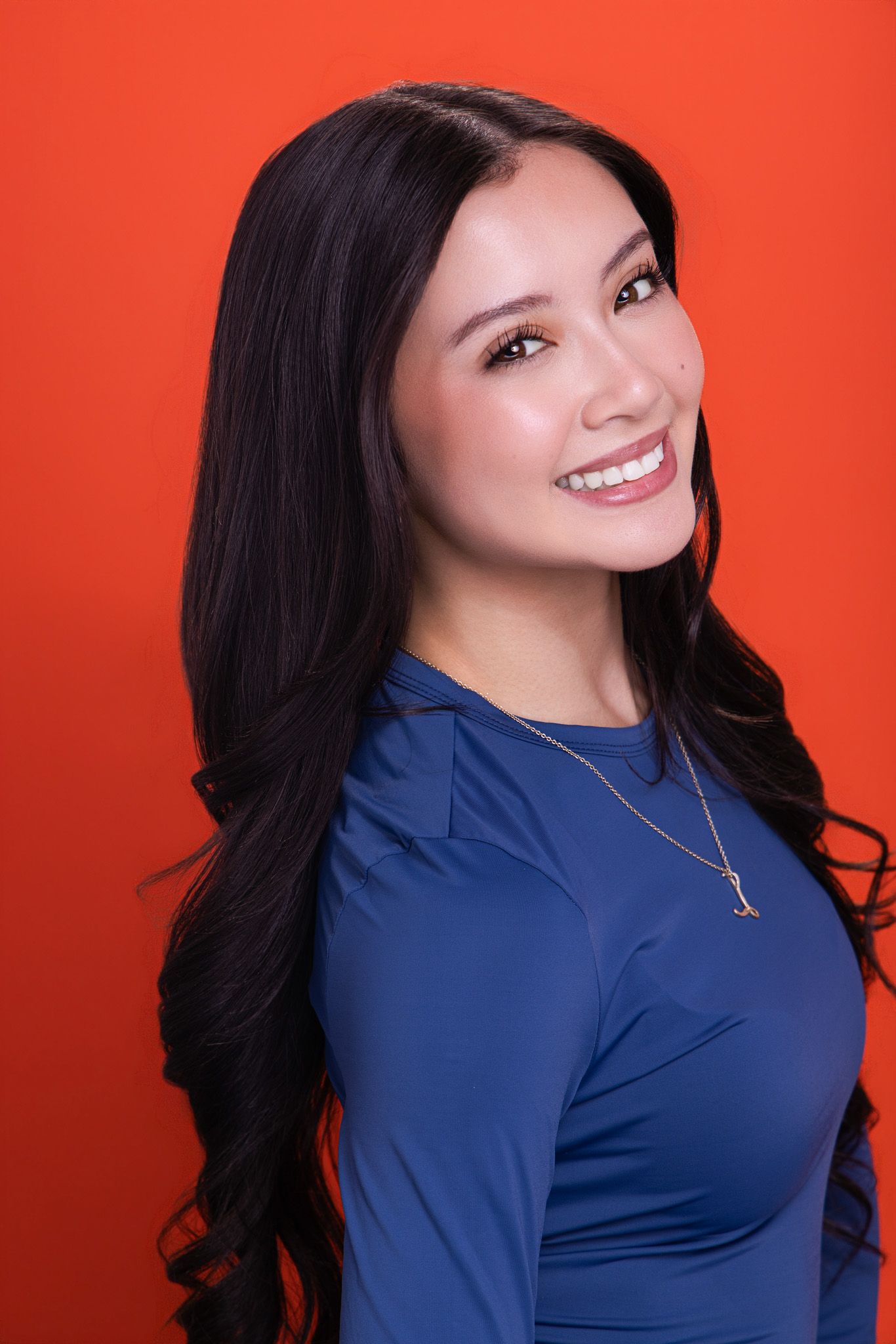 Smiling woman with long dark hair in blue long-sleeve top wearing gold necklace on red background.