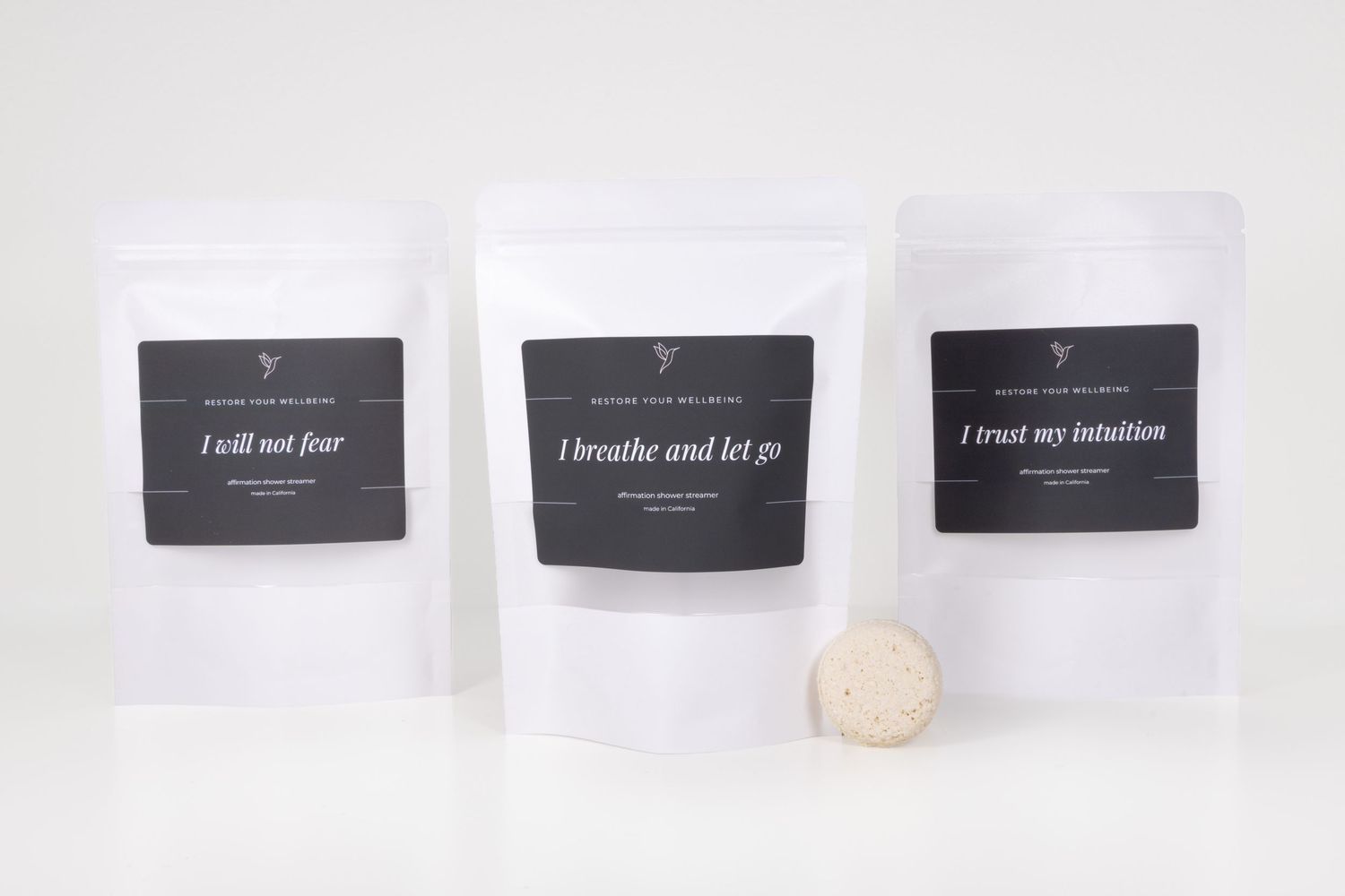 Three white kraft pouches with affirmation labels and a bath bomb for natural self-care products.