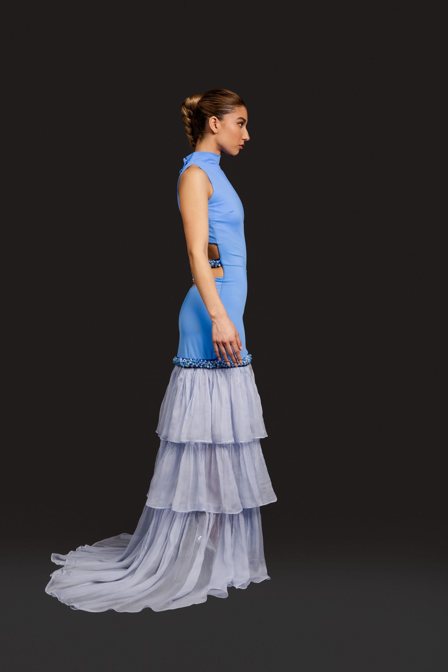 Woman in elegant blue sleeveless gown with tiered white tulle skirt and train, posing sideways against dark background.