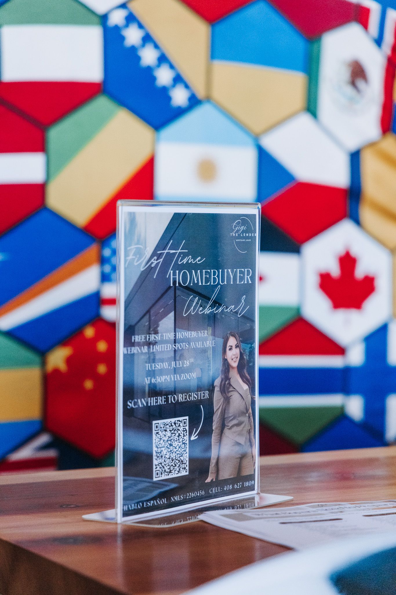 First-time homebuyer webinar flyer in acrylic stand on table with colorful international flags wall backdrop.