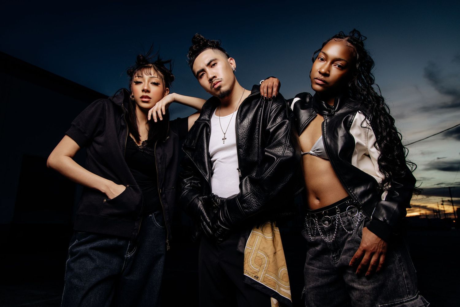 Three people in edgy black leather outfits pose together against a dramatic sunset sky backdrop.