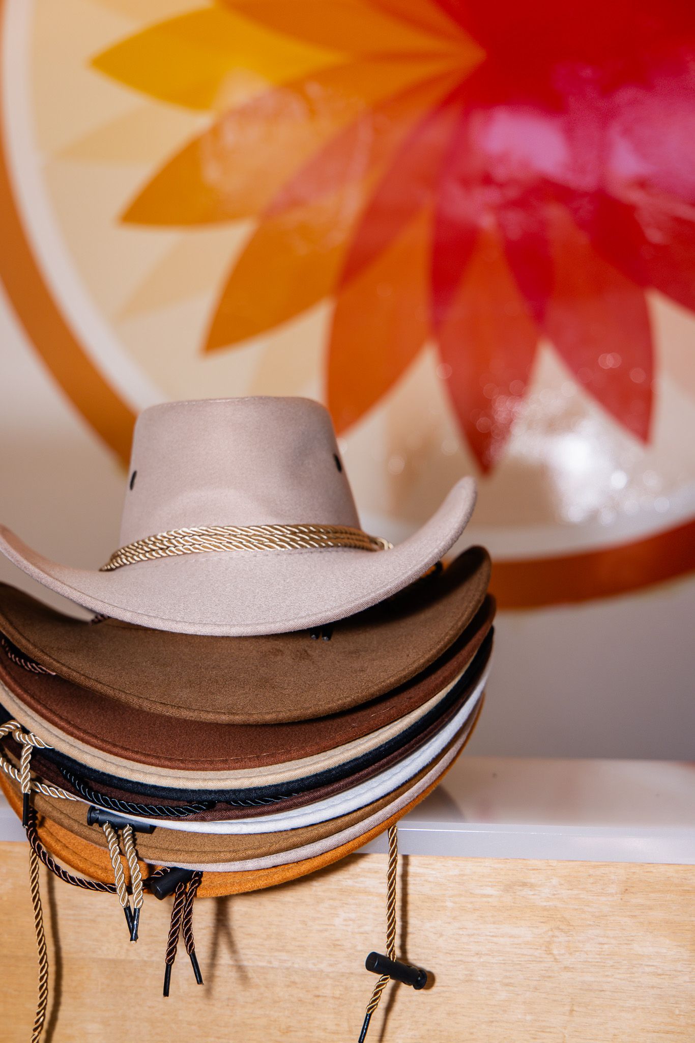 Stack of cowboy hats in various shades of brown and white with braided hat bands against blurred orange background.