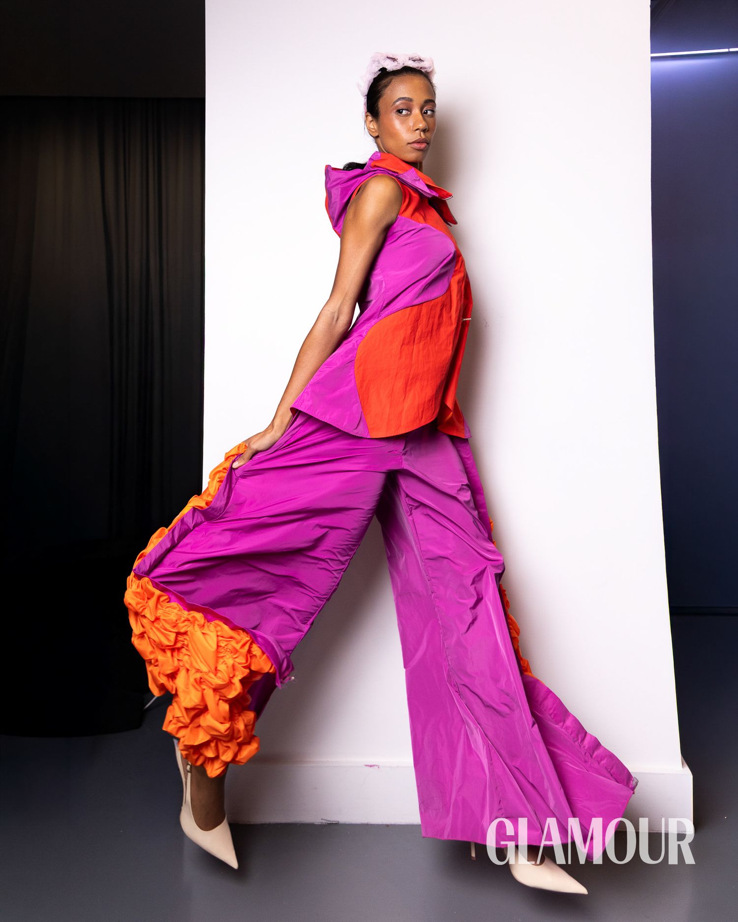 Fashion model poses in vibrant purple and orange flowing gown against dark backdrop creating dramatic movement effect.