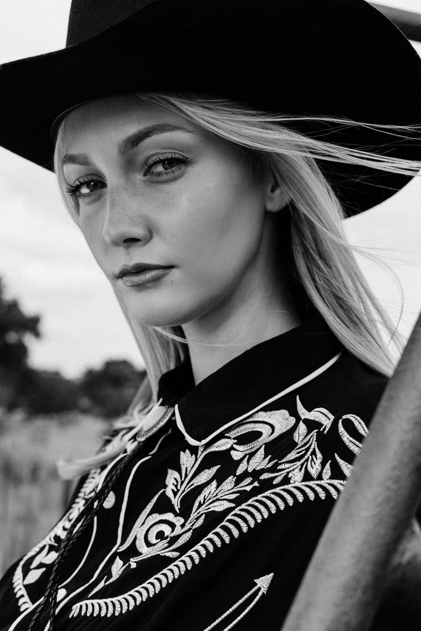 Artistic black and white portrait of a model in a western hat and embroidered shirt looking to the side.