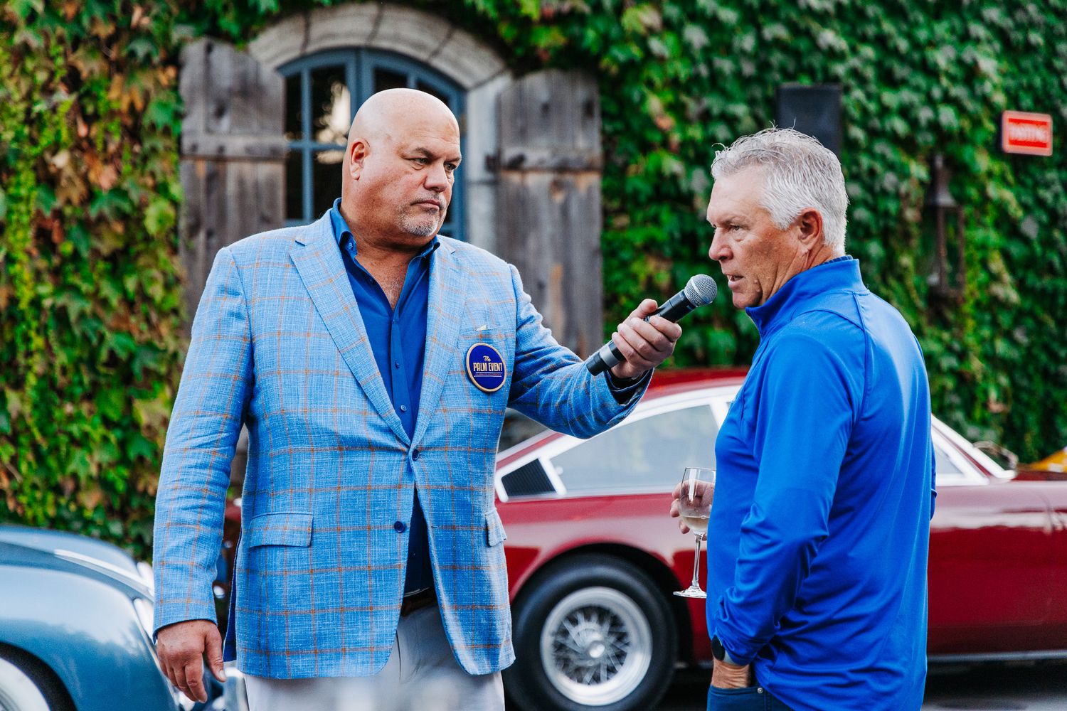 Two men in blue attire converse and laugh together outdoors against an ivy-covered wall background.