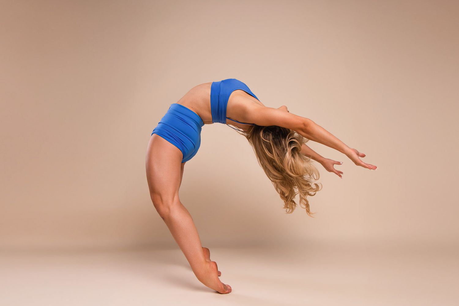 Woman in blue athletic wear performing a dramatic backbend stretch against a neutral beige studio background.