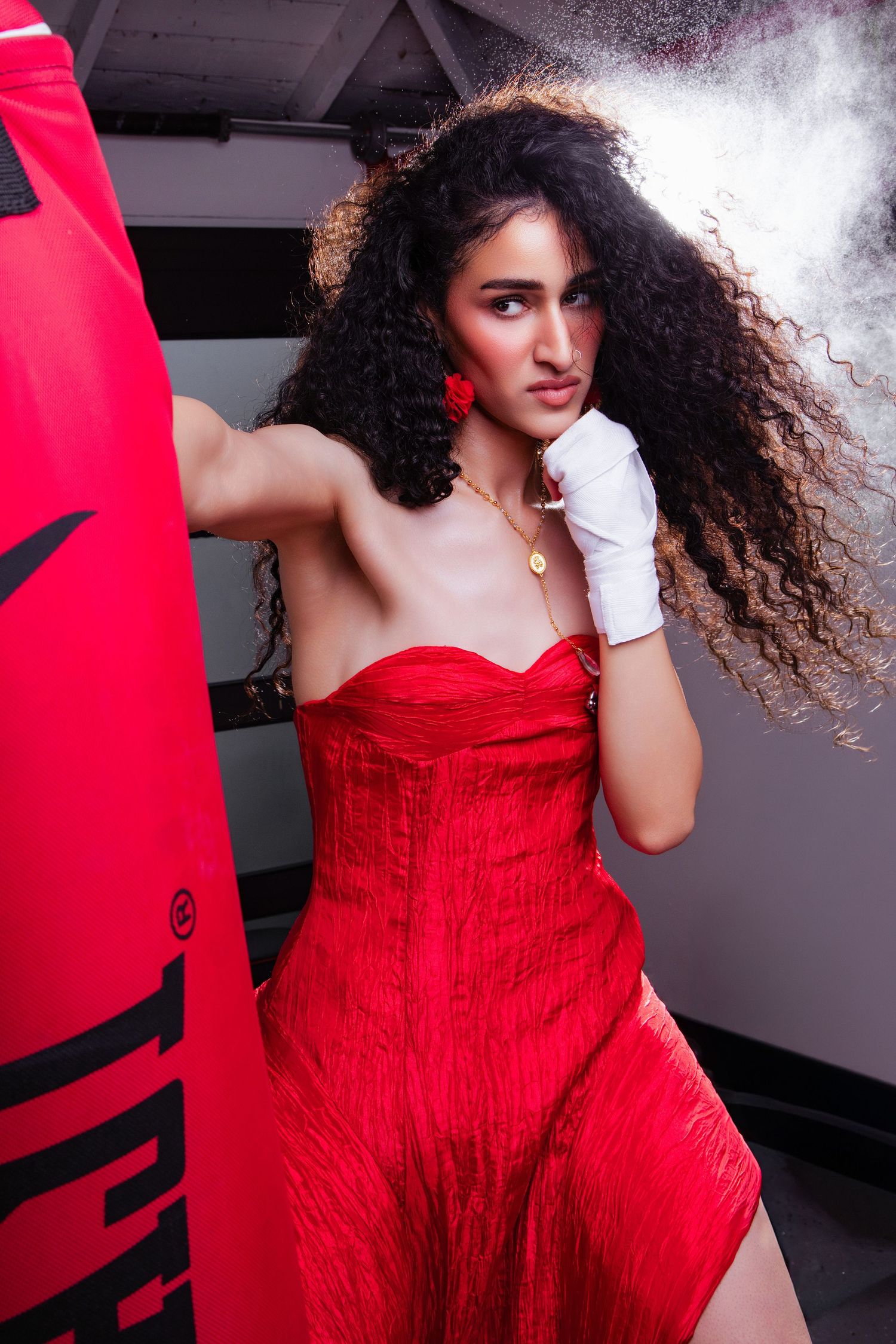 Woman in red strapless dress with white boxing wraps punches a red punching bag, curly hair flowing.