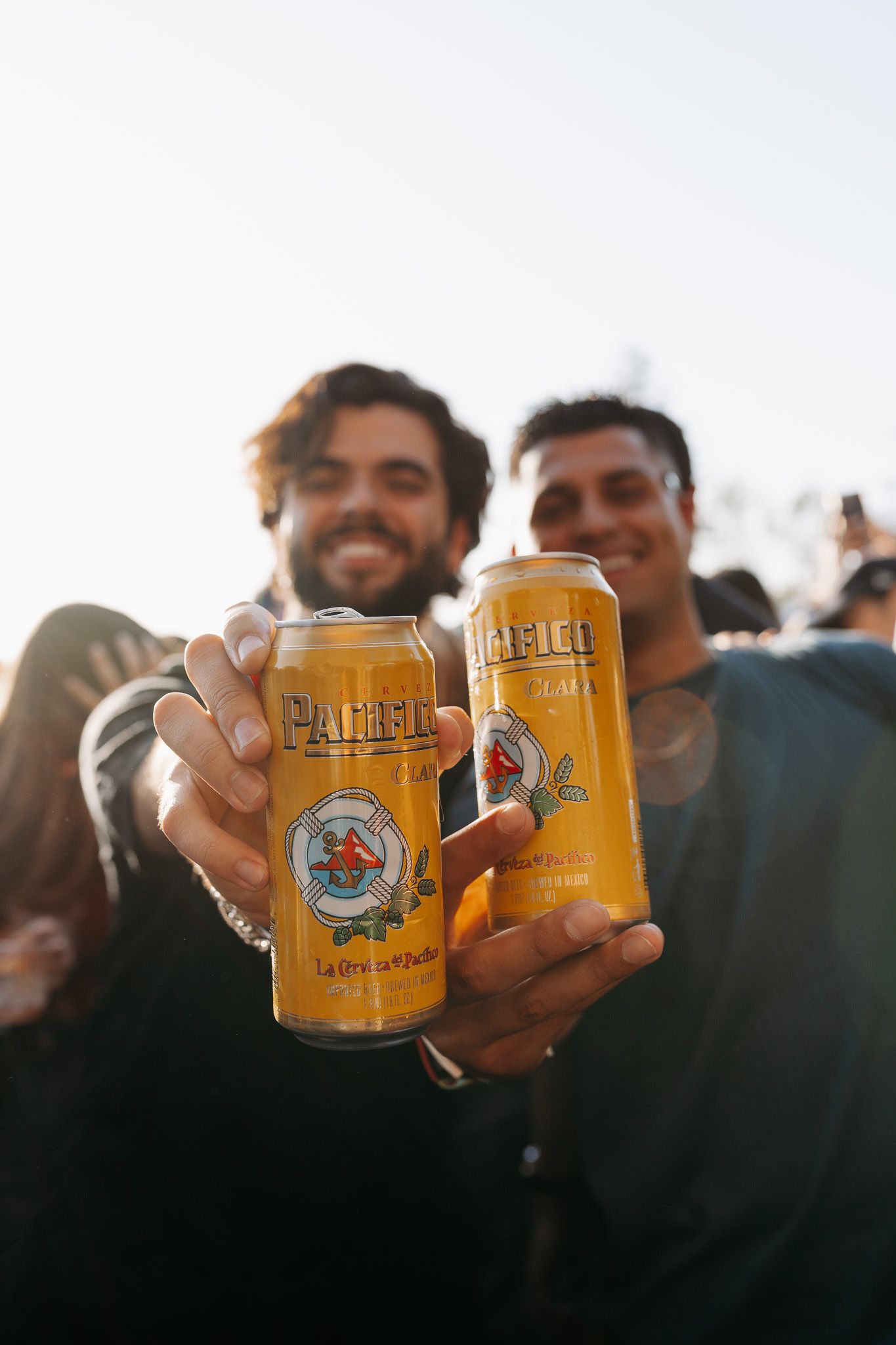 Two smiling men toasting with Pacifico beer cans outdoors at a sunny event.