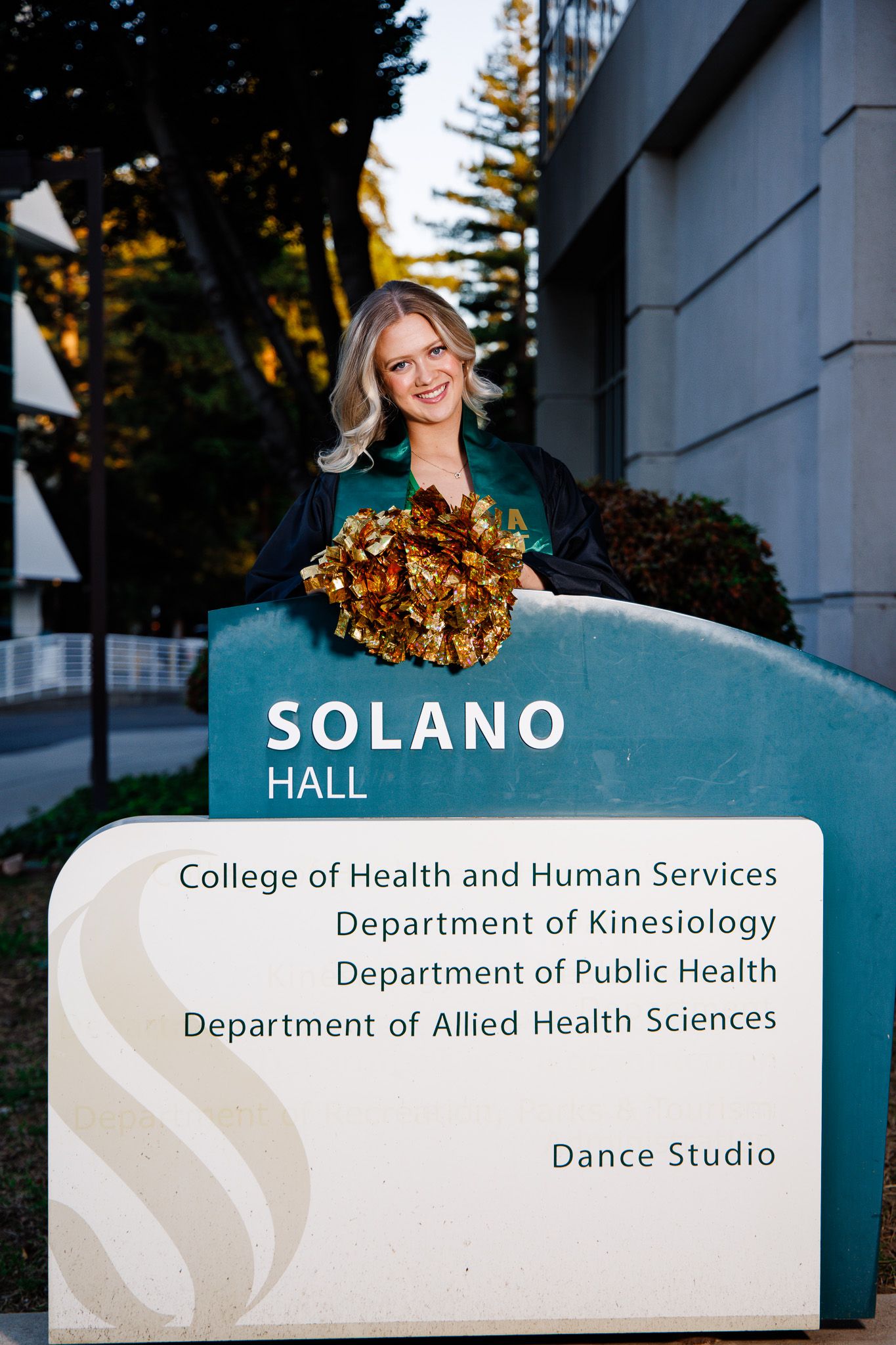 Graduate in cap and gown poses proudly at Solano Hall sign at College of Health and Human Services.
