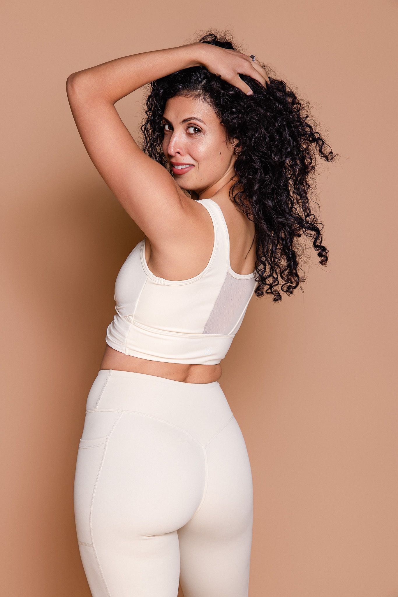 Woman in white activewear set posing from behind showing curly hair in beige studio.