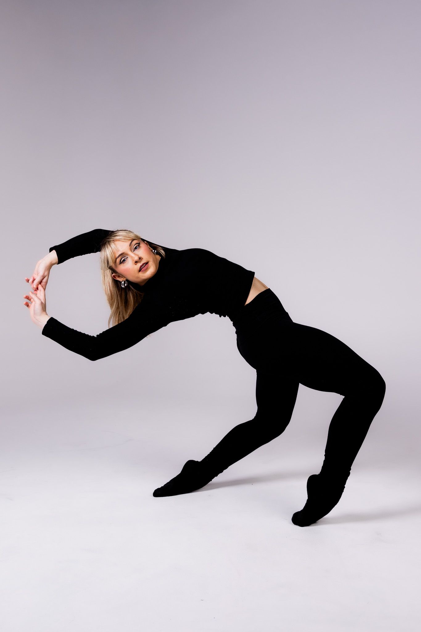 A dancer in all black attire performs a graceful bending pose against a light gray studio background.
