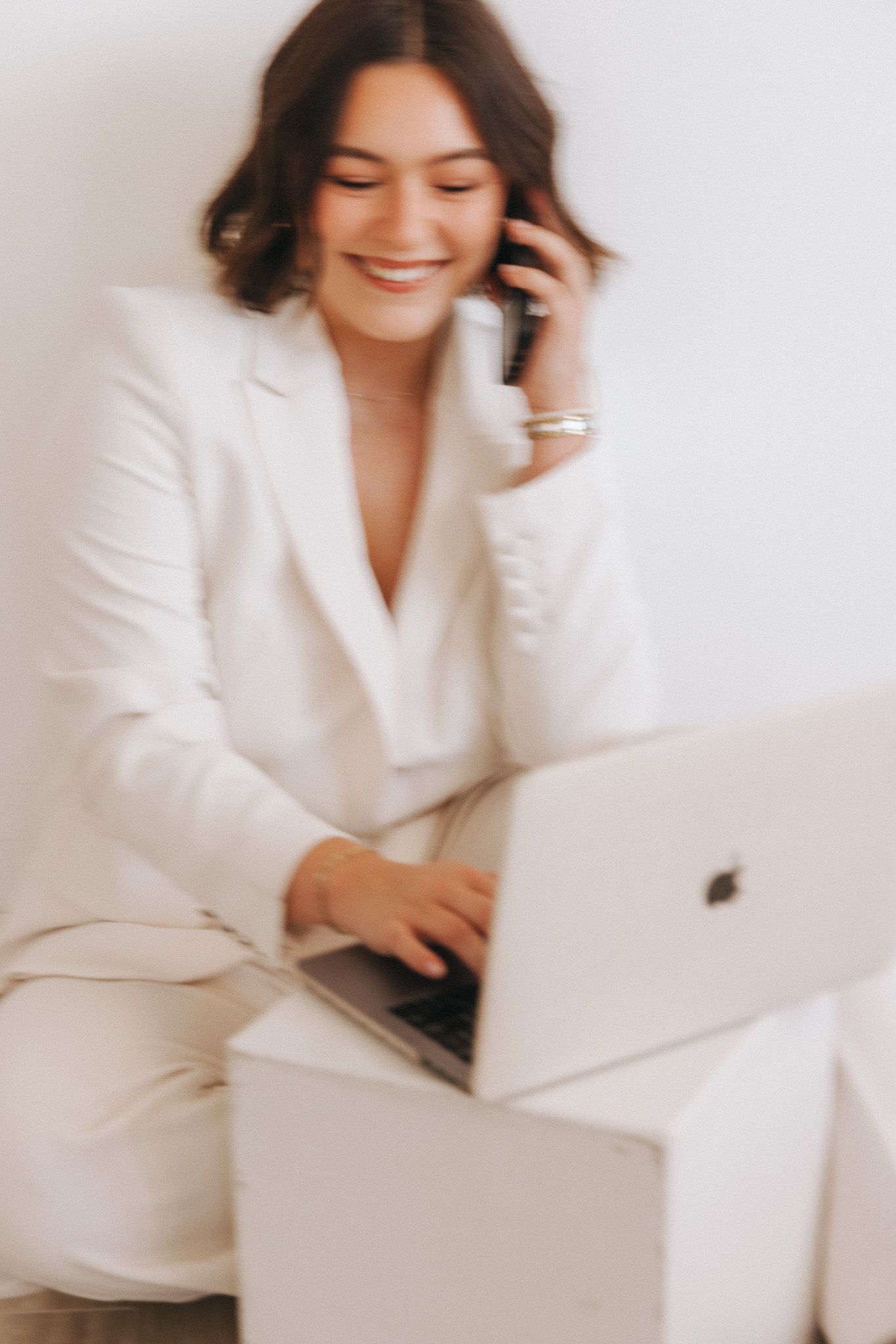 Professional in a white blazer works on a MacBook laptop while making a phone call.