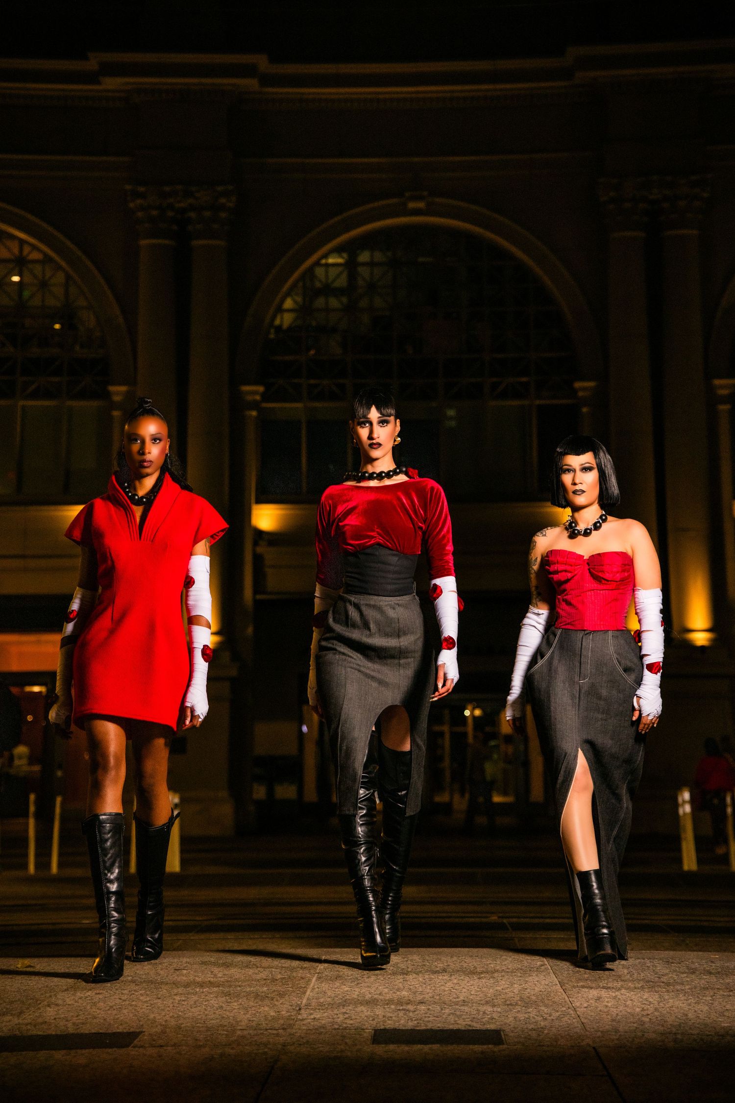 Models in red tops and black boots pose in dramatic lighting against a dark urban backdrop during a fashion shoot.