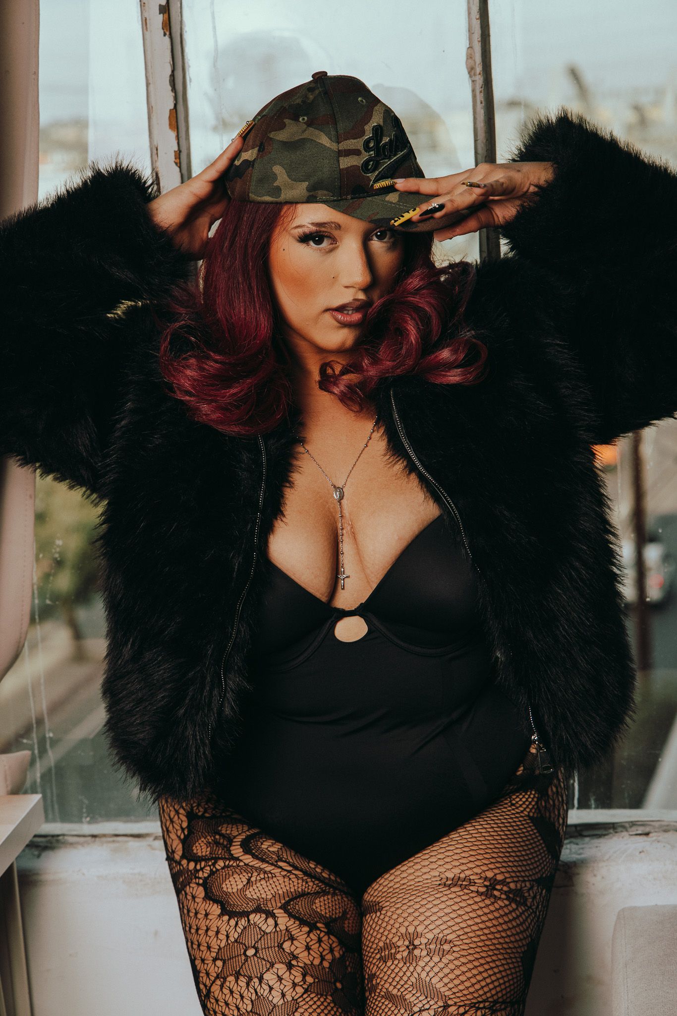 Model in black bodysuit and fur coat poses wearing camouflage cap and fishnet stockings in moody lighting.