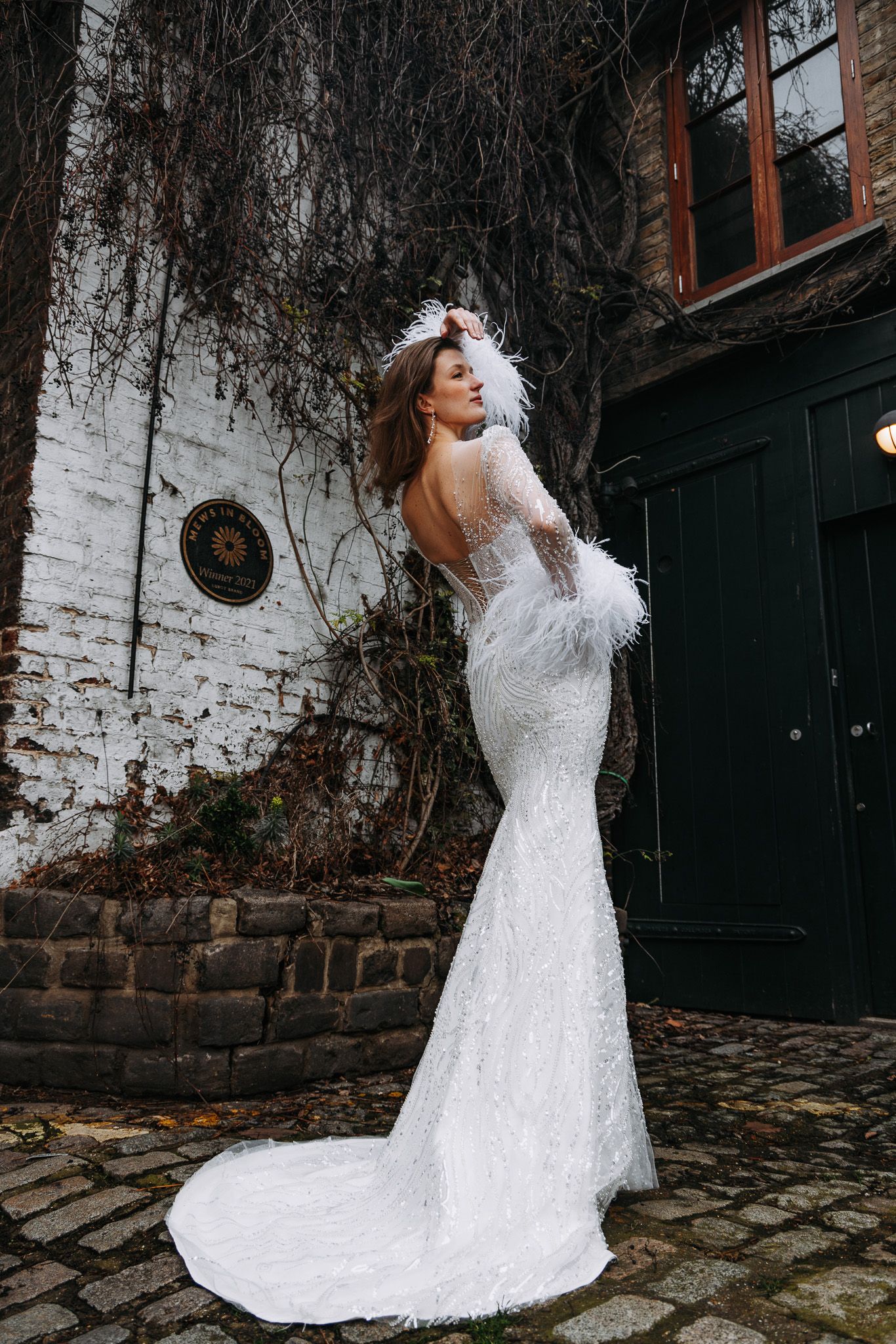 Elegant white lace mermaid wedding gown with feather details and open back shown against rustic brick wall backdrop.