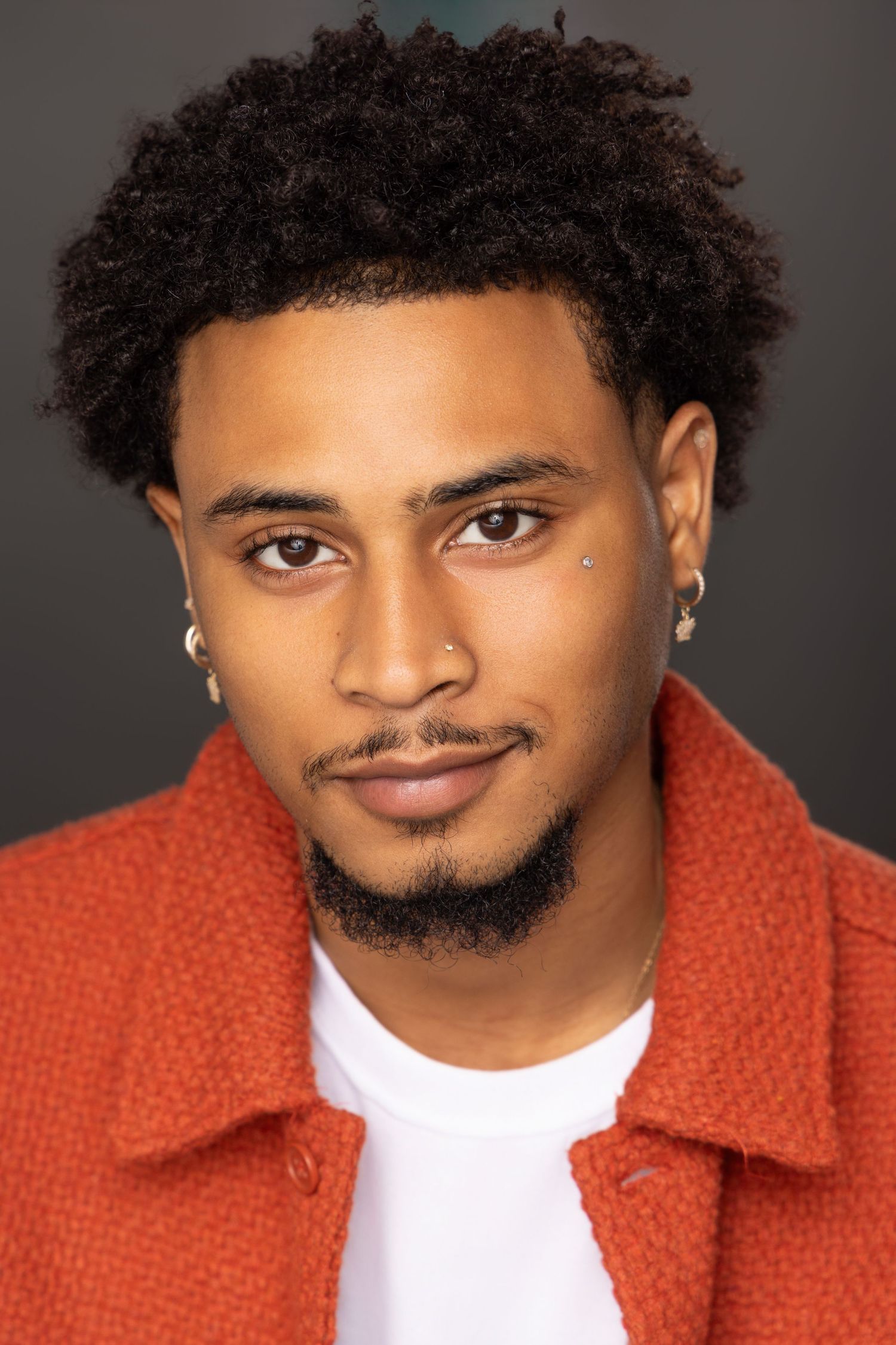 A person in a bright orange jacket and white t-shirt poses against a dark background for a professional headshot.