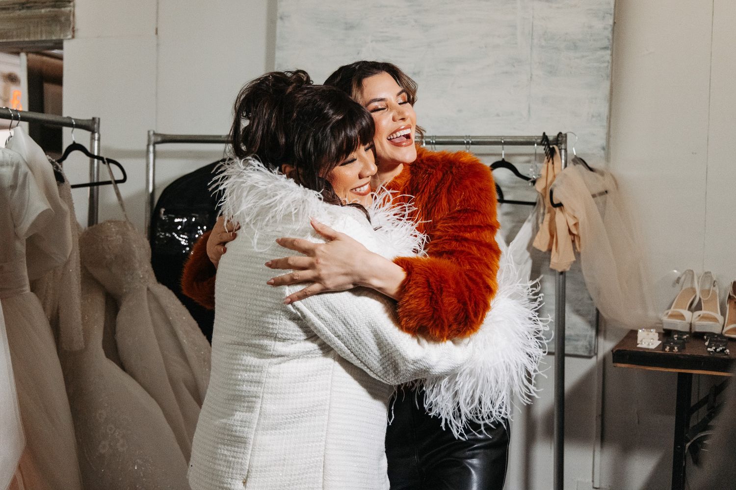 Two people share a joyful embrace in a bridal boutique surrounded by clothing racks and shoes.