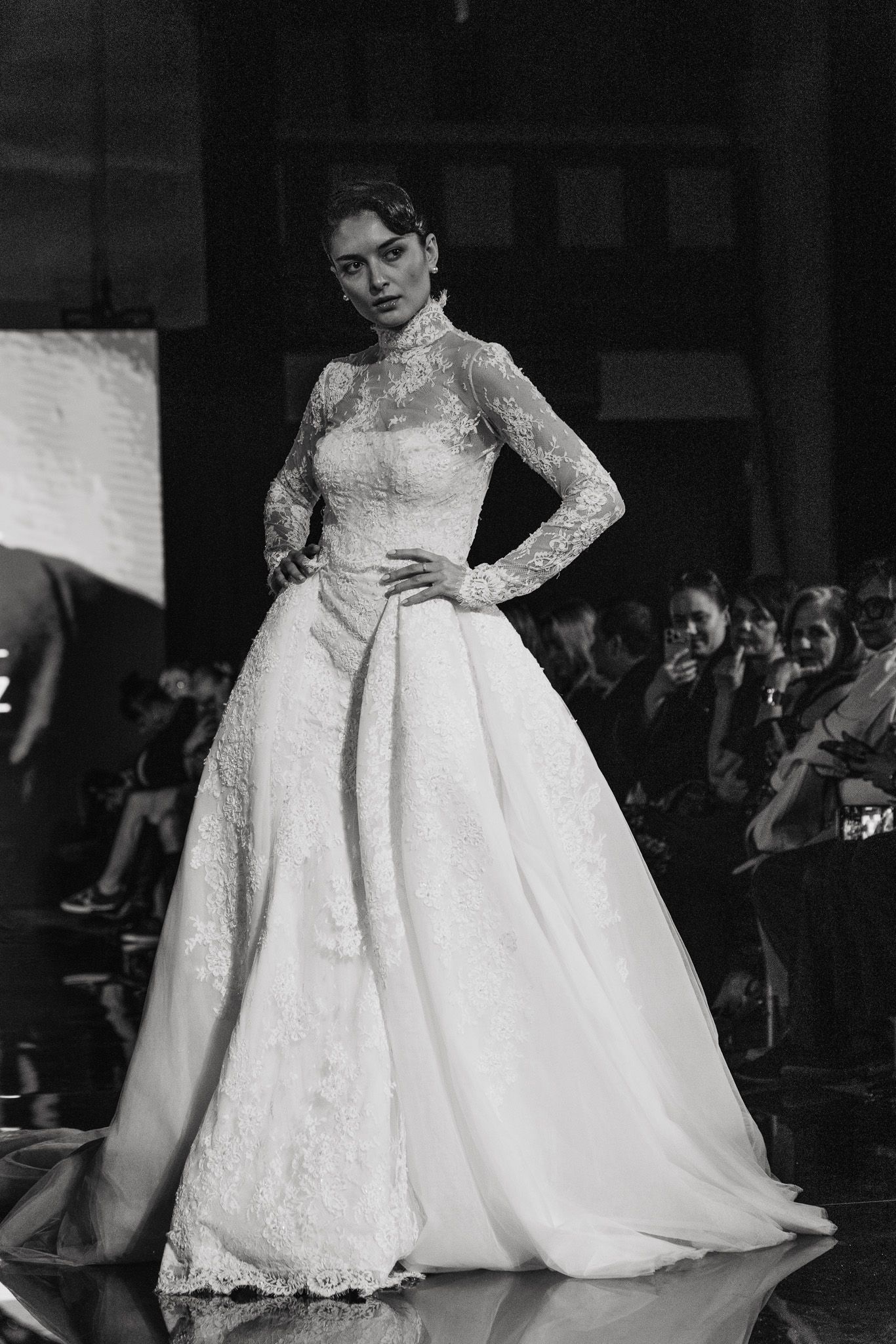 A model walks the runway in an elegant white lace long-sleeved wedding gown with a full skirt at a bridal fashion show.