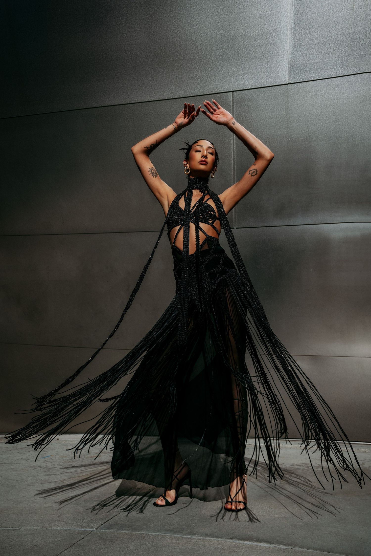 Woman in black fringed halter gown poses dramatically with arms raised against metallic wall.