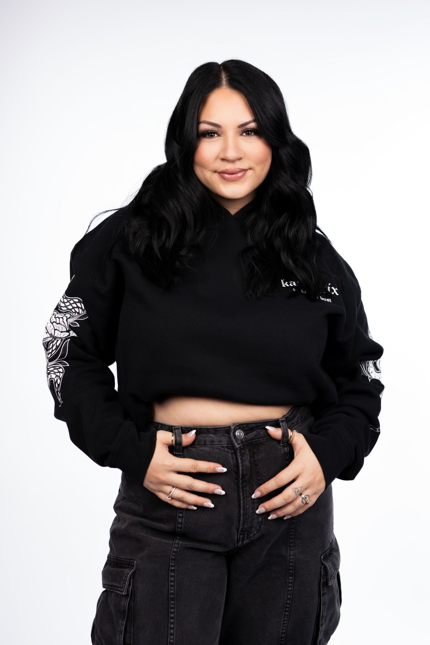 Woman in black cropped hoodie with graphic sleeve detail, paired with black cargo jeans, on white background.
