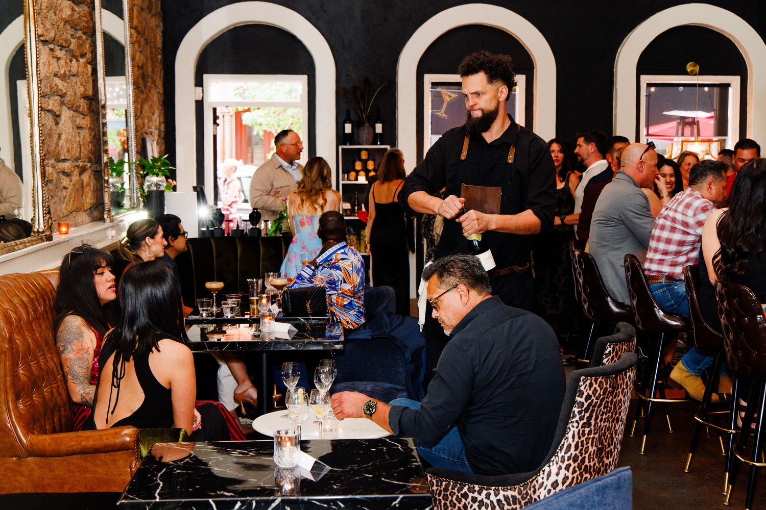 Busy restaurant interior with waiter serving guests at marble tables in an elegant venue with arched black walls.
