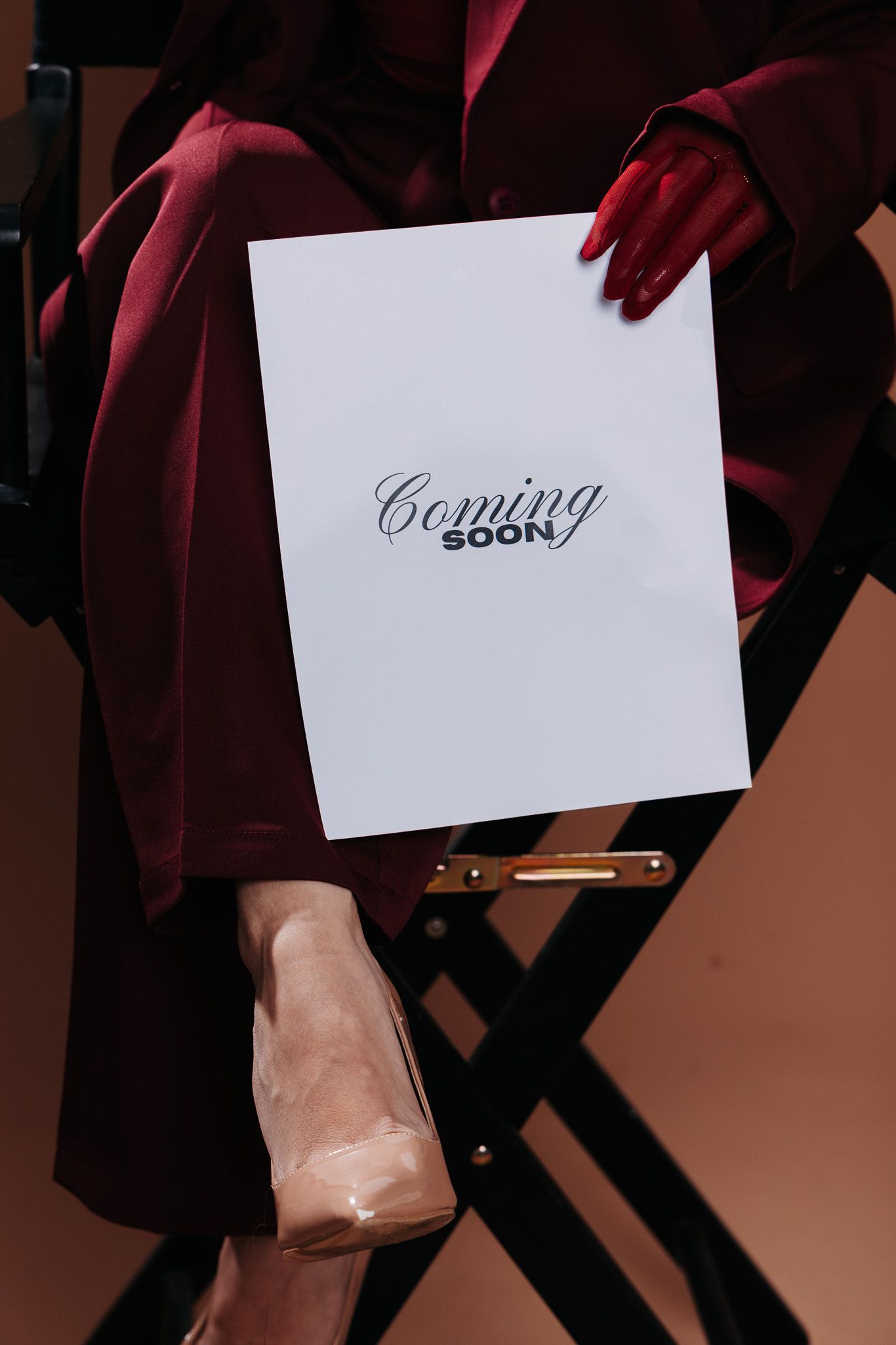 Woman in burgundy outfit sitting in director's chair holding a white 'Coming Soon' sign with red manicured nails.