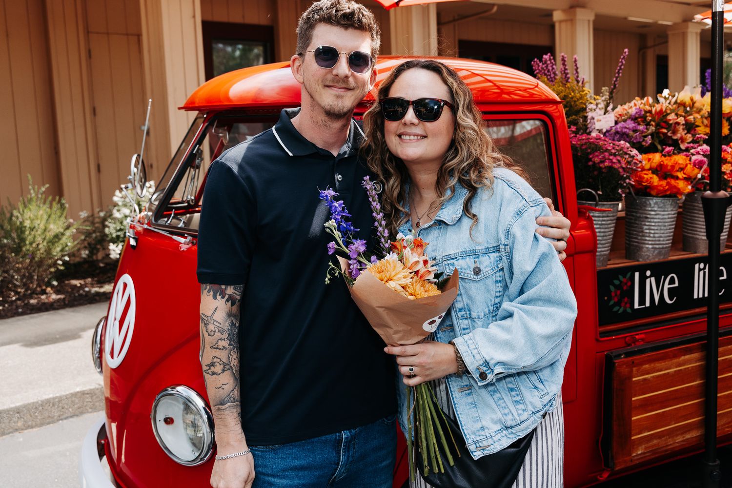 Couple poses smiling in front of red VW van with colorful flowers at outdoor market.