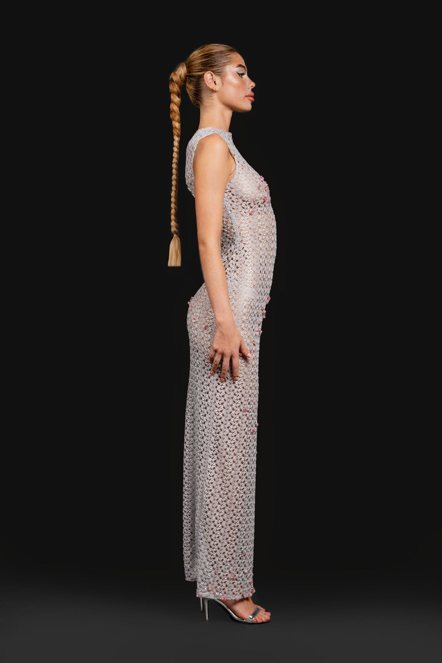 Side profile of model in sleeveless silver crochet maxi dress with long braided ponytail and strappy heels.