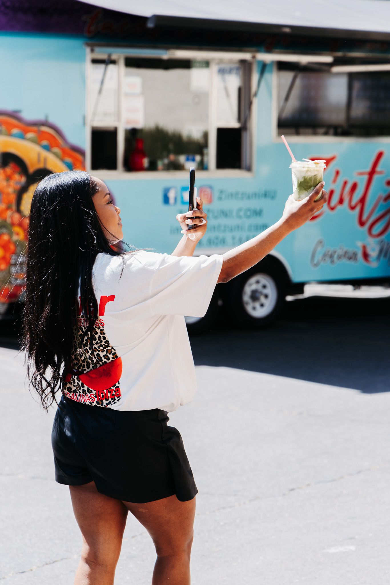 Someone with long dark hair wearing a white t-shirt and black shorts holds up a drink in front of a food truck.